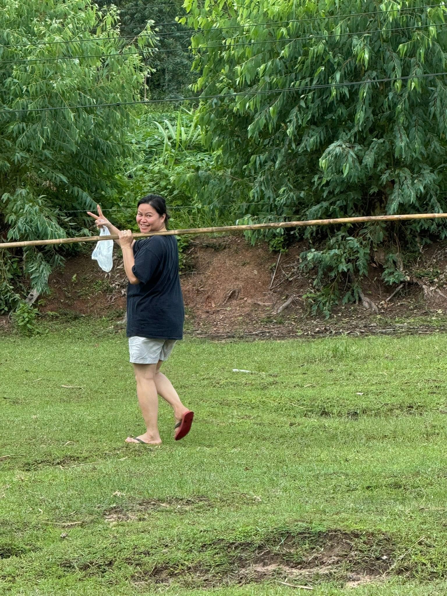 Jiratchaya carrying a long bamboo harvesting pole across her shoulder, smiling and walking through the green grounds of the Magic Land